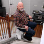 Man seated on the Bruno Elan stair lift at the bottom of the stairs, using the hand control