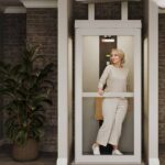 Harmar Spire Home Elevator with a full-height glass door and a folded tip-up seat, a woman standing inside the cabin